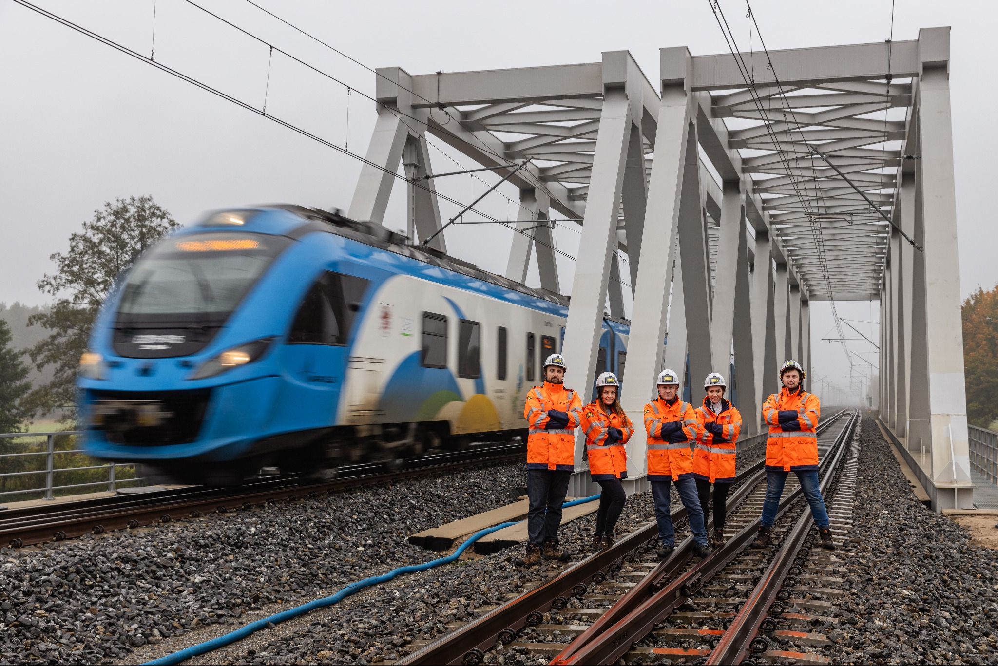Grupa inżynierów w pomarańczowych kurtkach i kaskach stoi na torach kolejowych na moście, w tle przejeżdża niebieski pociąg, widoczne elementy konstrukcji mostu i szyny.