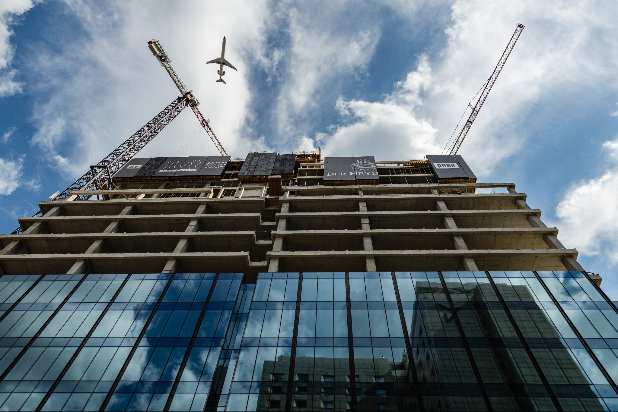 Niski kąt ujęcia szkieletu budynku w trakcie budowy, z żurawiami i przelatującym samolotem na tle błękitnego nieba z chmurami, odbicie budynku w szklanej fasadzie.