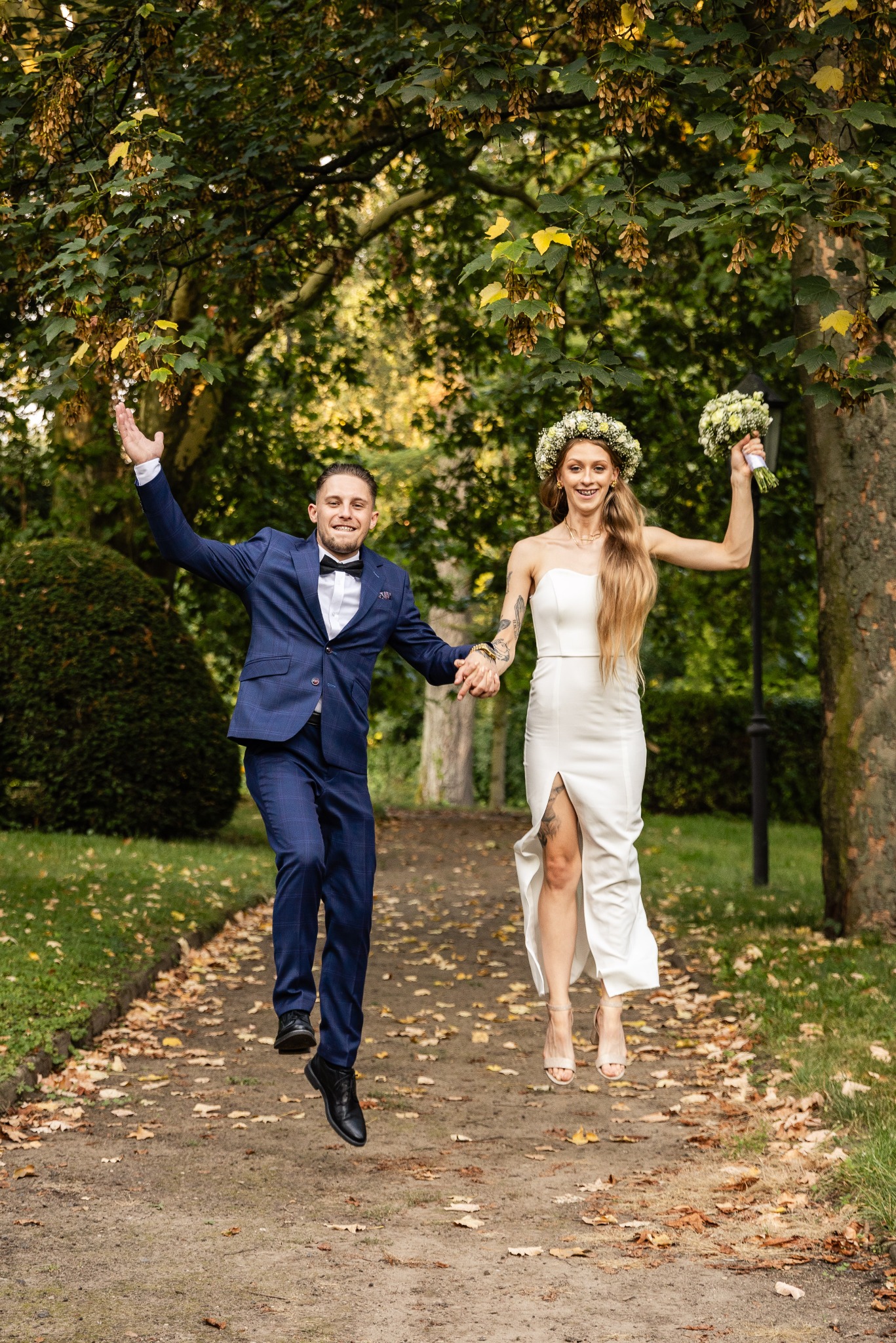 Para młoda w strojach ślubnych skacze radośnie na ścieżce w parku, panna młoda w białej sukni z rozcięciem i wiankiem na głowie, pan młody w niebieskim garniturze w kratę.