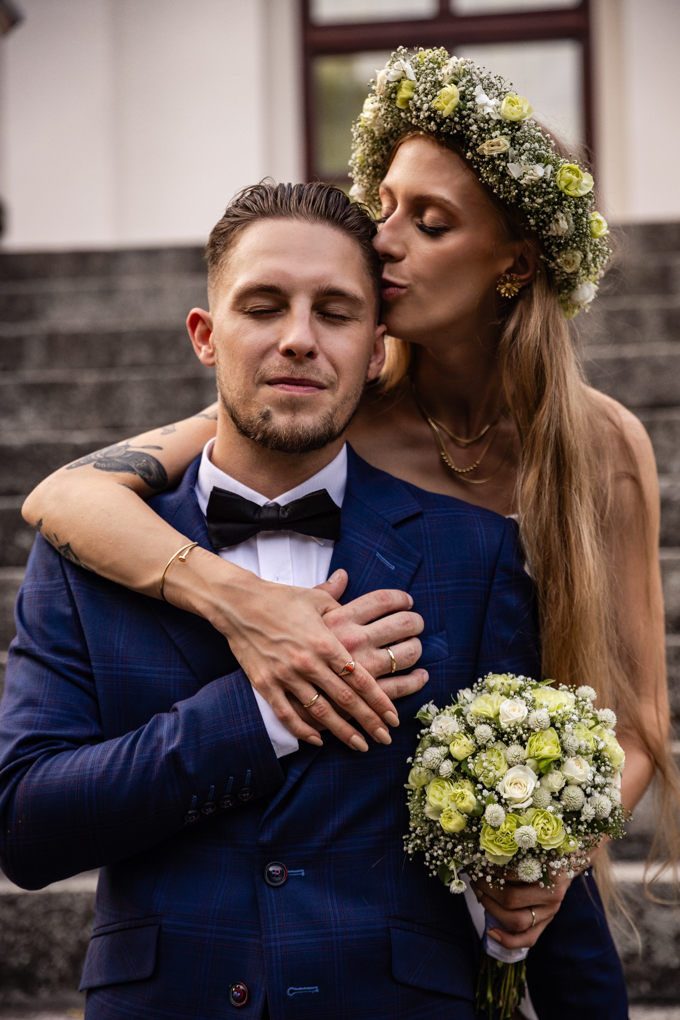 Panna młoda w wianku z białych i jasnozielonych kwiatów całuje w policzek pana młodego w granatowym garniturze w kratę, bukiet ślubny z białych i zielonych kwiatów w dłoniach, na tle kamiennych...