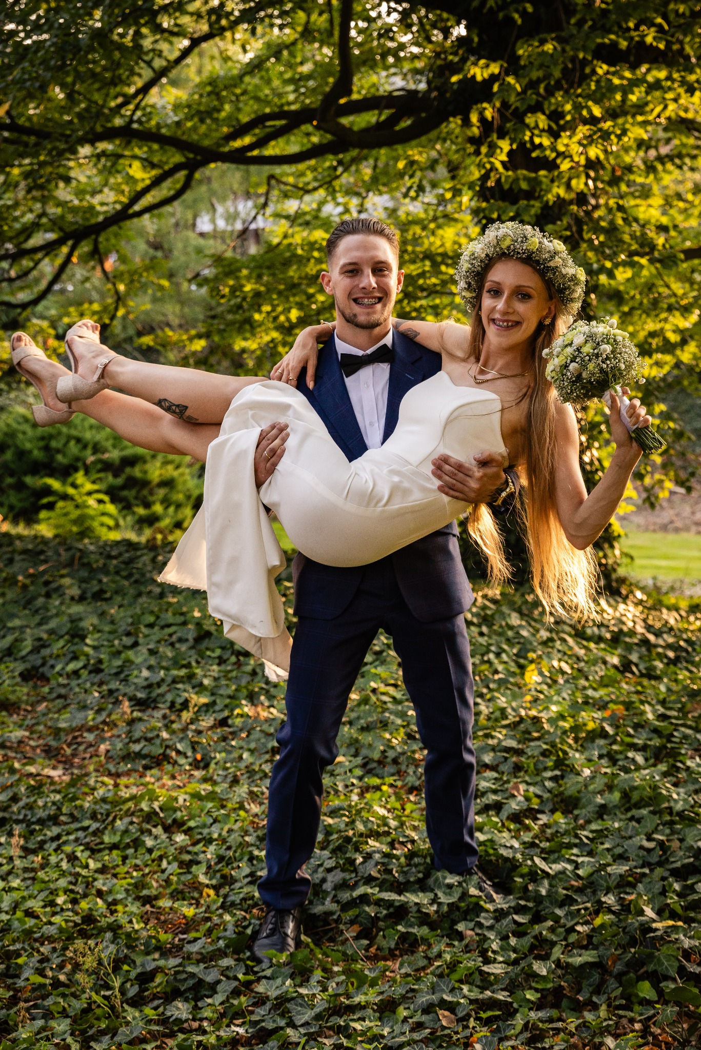 Fotograf z Szamotuł uchwycił moment radości: Pan młody trzyma na rękach uśmiechniętą pannę młodą w wianku i z bukietem na tle zieleni.