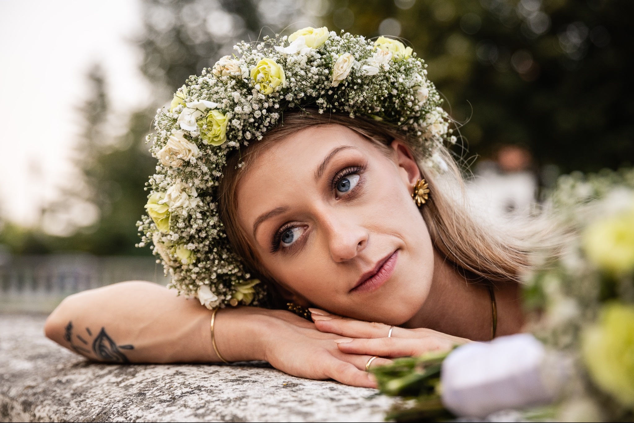Portret kobiety z wiankiem z białych i żółtych kwiatów na głowie, opierającej się na kamiennym murku, z tatuażem na przedramieniu i złotymi kolczykami w kształcie kwiatów.