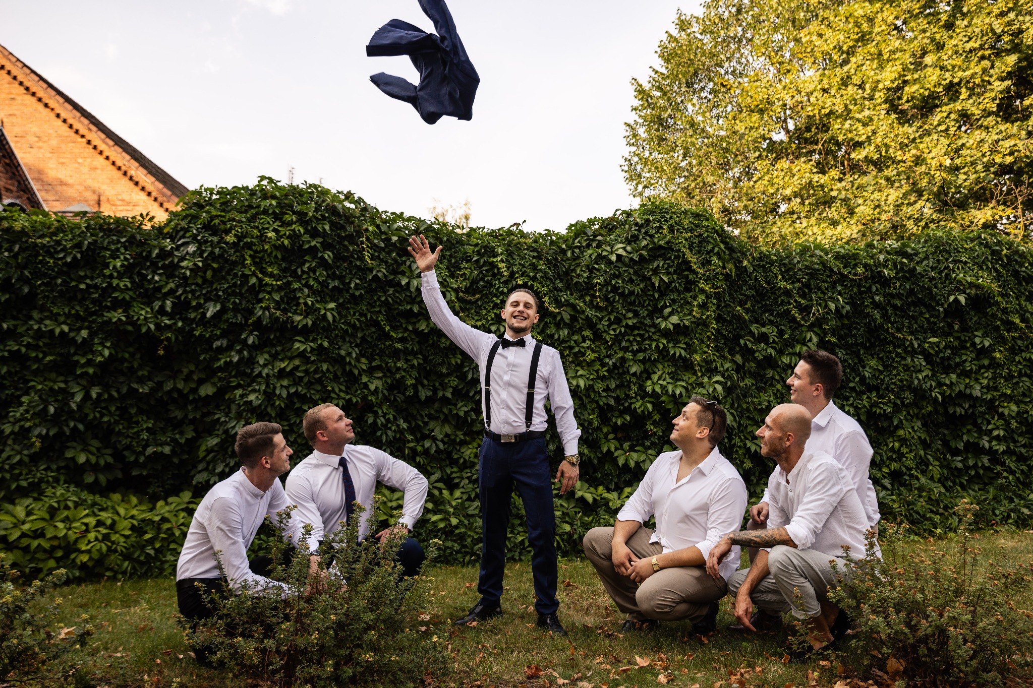 Pan młody z druhami świętuje rzucając marynarkę w górę na tle zielonej ściany z bluszczu i ceglanego budynku.