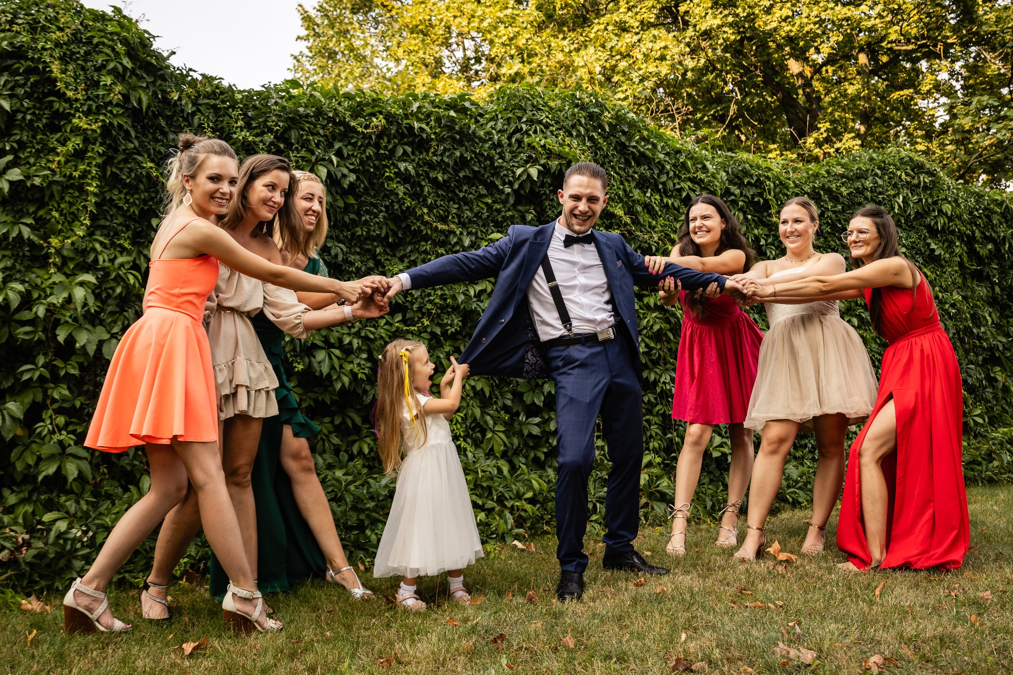 Uśmiechnięty pan młody w granatowym garniturze z szelkami, otoczony druhnami w kolorowych sukienkach, trzymającymi się za ręce; mała dziewczynka w białej sukience ciągnie go za marynarkę na tle...