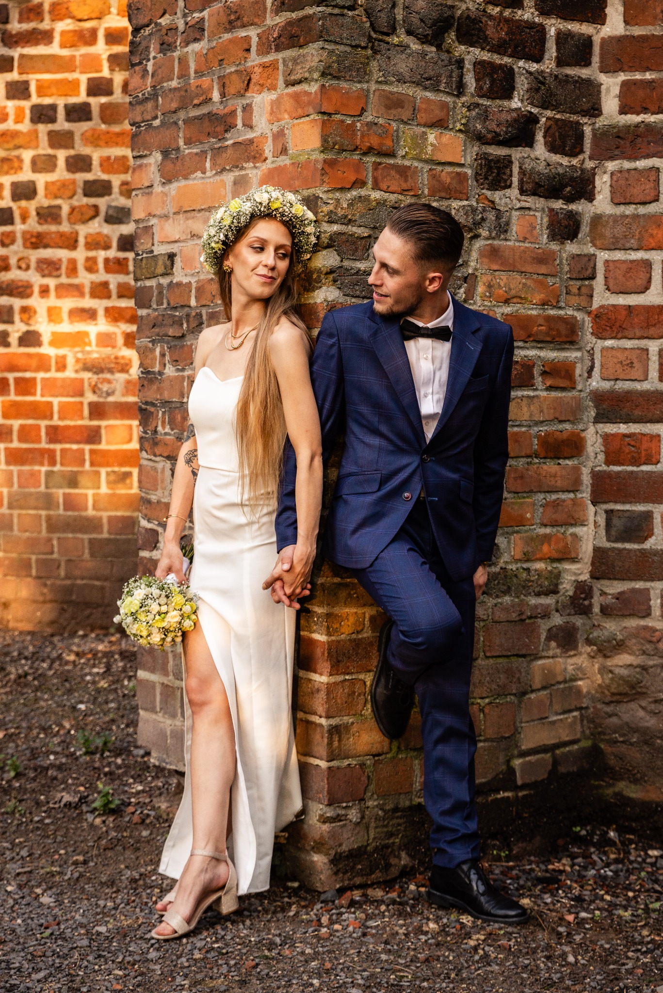 Para młoda, on w granatowym garniturze w kratę, ona w prostej sukni z rozcięciem i wiankiem, trzymają się za ręce, opierając się o ceglany mur.