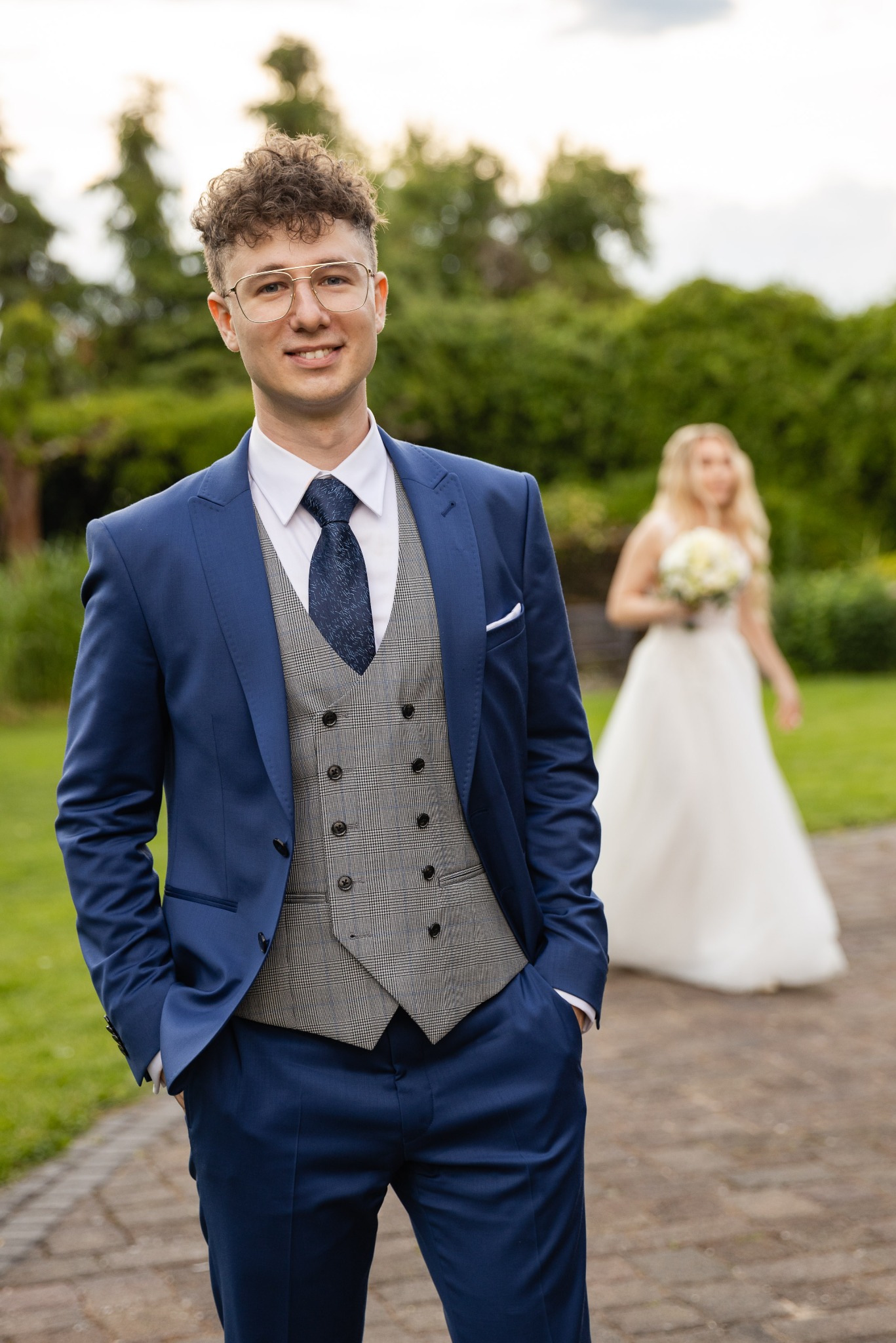 Uśmiechnięty pan młody w niebieskim garniturze i wzorzystej kamizelce, z narzeczoną w tle, trzymającą bukiet. Plenerowa sesja ślubna w parku.