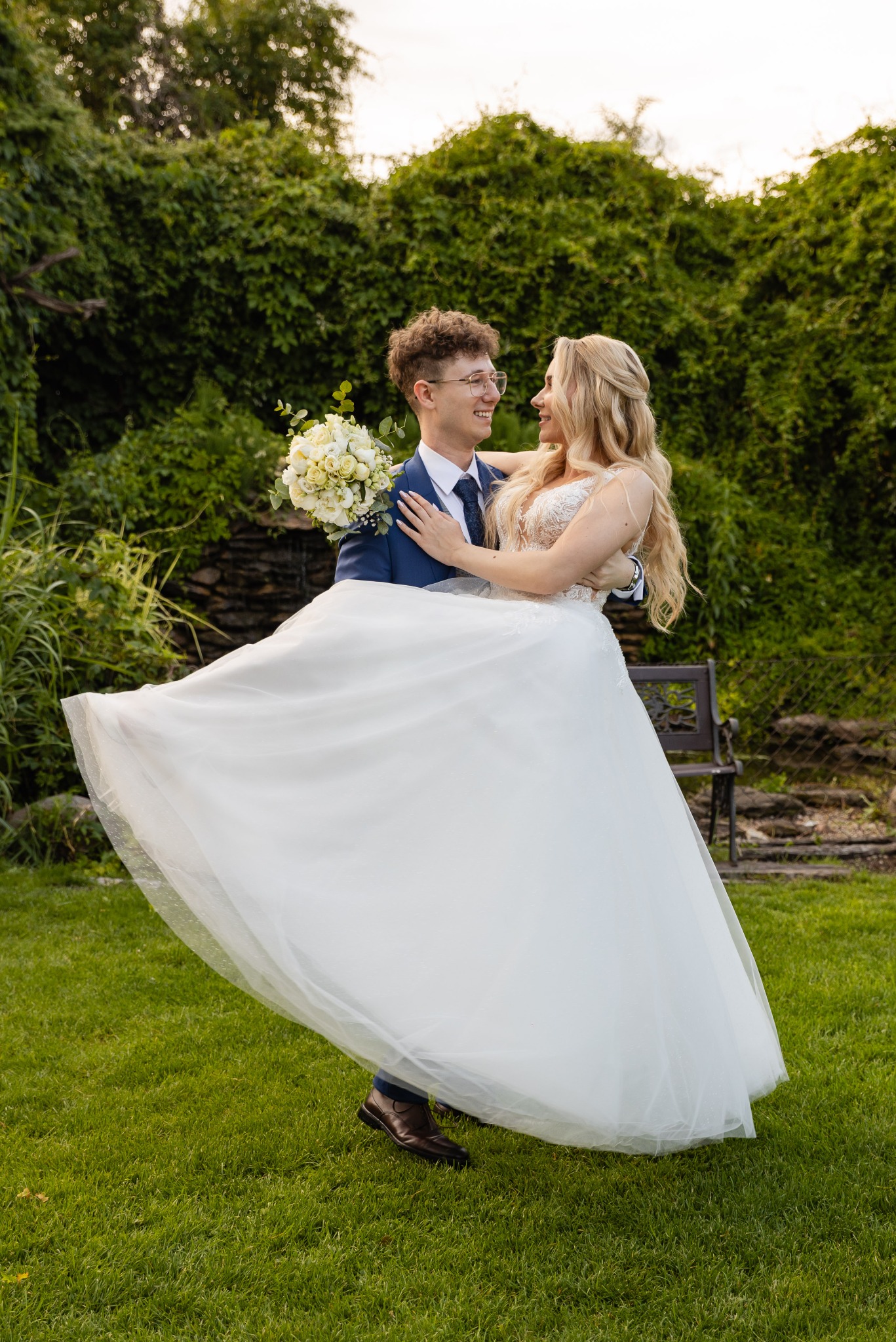 Uśmiechnięty pan młody w niebieskim garniturze trzyma na rękach pannę młodą w białej sukni z bukietem na tle zieleni i małego wodospadu.