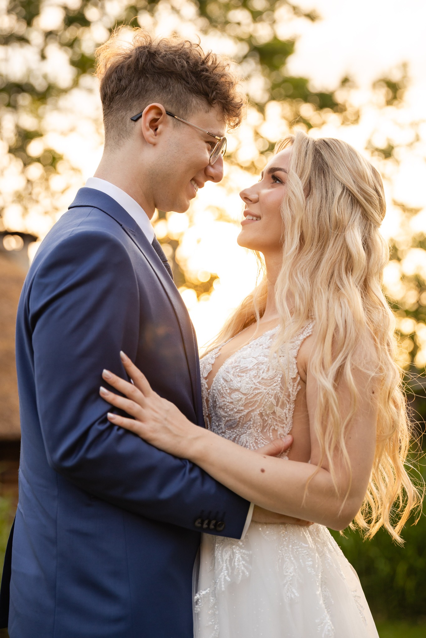 Uśmiechnięta para młoda w objęciach, on w granatowym garniturze i okularach, ona w koronkowej sukni, w tle rozmyte, złote światło zachodzącego słońca.