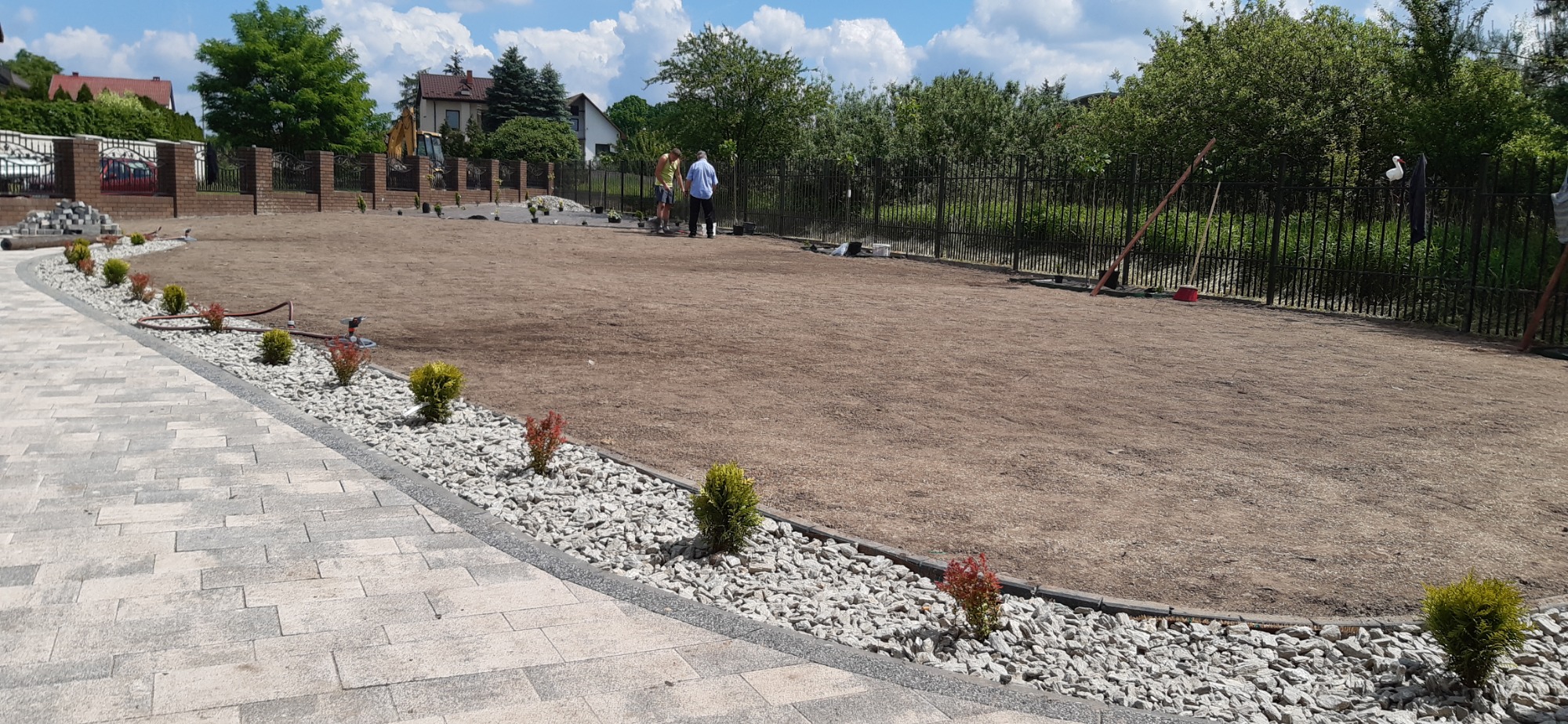 Przygotowanie terenu pod trawnik: wyrównana ziemia, obrzeża z kostki i białego grysu, nasadzenia małych krzewów ozdobnych, widoczni dwaj mężczyźni przy pracy.