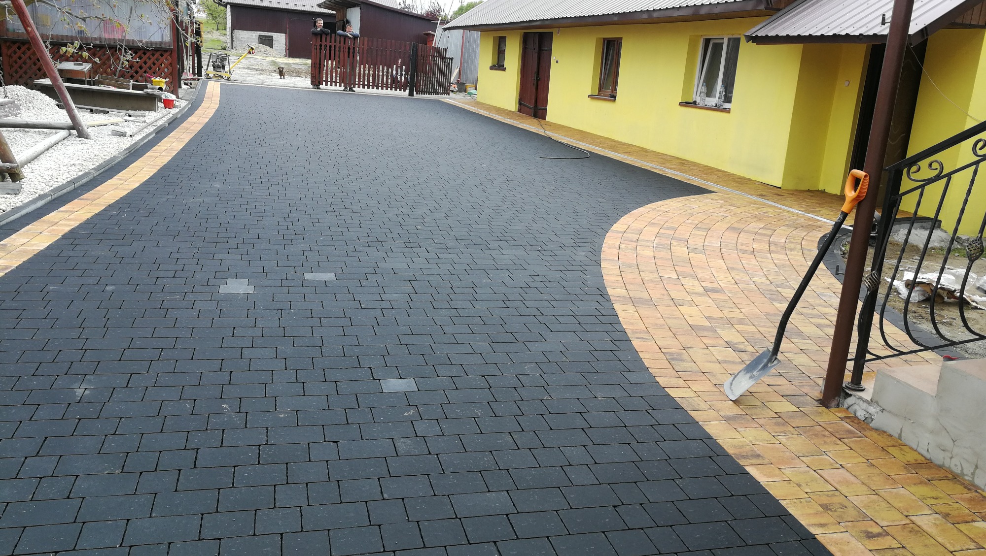 Ułożona nawierzchnia z ciemnej kostki brukowej z obramowaniem z kostki w odcieniach żółci, widoczna łopata oparta o metalową barierkę schodów wejściowych do żółtego budynku.