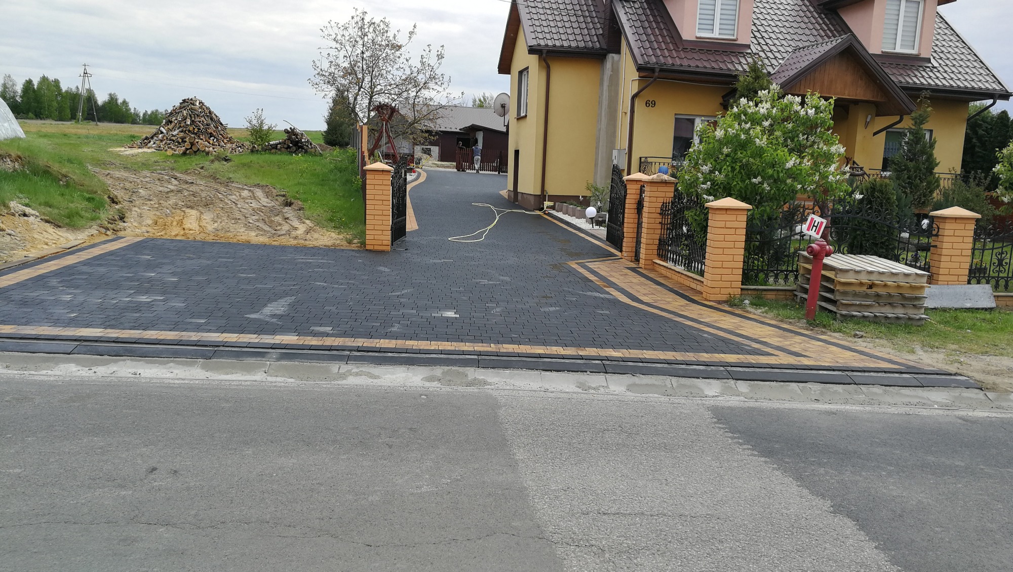 Ułożona ciemnoszara kostka brukowa z obramowaniem w kolorze piaskowym tworząca podjazd do domu z bramą z ceglanymi słupkami. W tle sterta drewna i budynek gospodarczy.