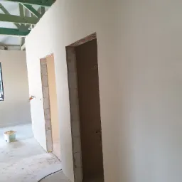 Świeżo położone gładzie na ścianach wewnętrznych budynku w trakcie remontu, widoczne ościeżnice drzwiowe i surowy strop z drewnianych belek.