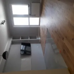 Wnętrze mieszkania w trakcie prac wykończeniowych: drewniana podłoga w jodełkę francuską, fragment szarej wylewki samopoziomującej, ściana z półkami, wiszący telewizor i lampa sufitowa.