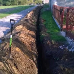 Długi wykop w ziemi z widoczną rurą na dnie, obok czerwone ogrodzenie i łopata z zielonym uchwytem. Ślad po wykopie ciągnie się wzdłuż drogi.