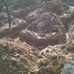 Przygotowanie wykopu pod fundament z ułożonym zbrojeniem. W tle sterta ziemi i fragment rury. Widok z góry na wykop.