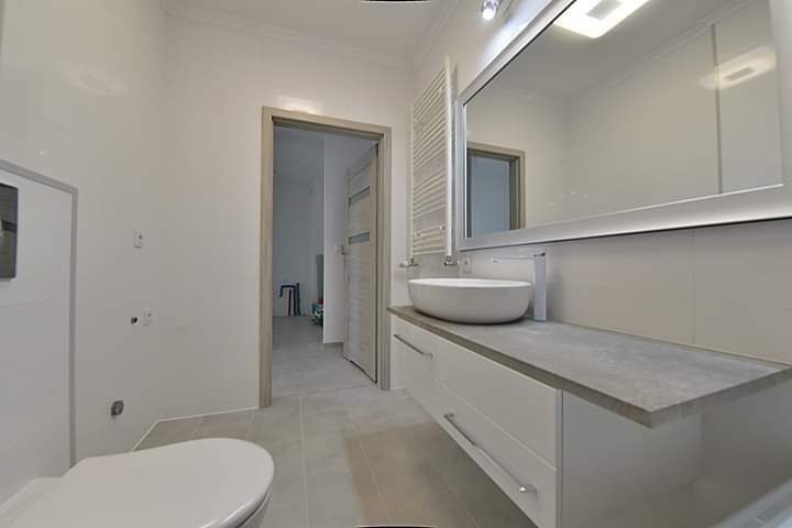 Nowoczesna łazienka z białymi płytkami, wiszącą umywalką na szafce z szufladami, dużym lustrem z oświetleniem LED i grzejnikiem drabinkowym, widoczny fragment toalety i drzwi do innego pomieszczenia.