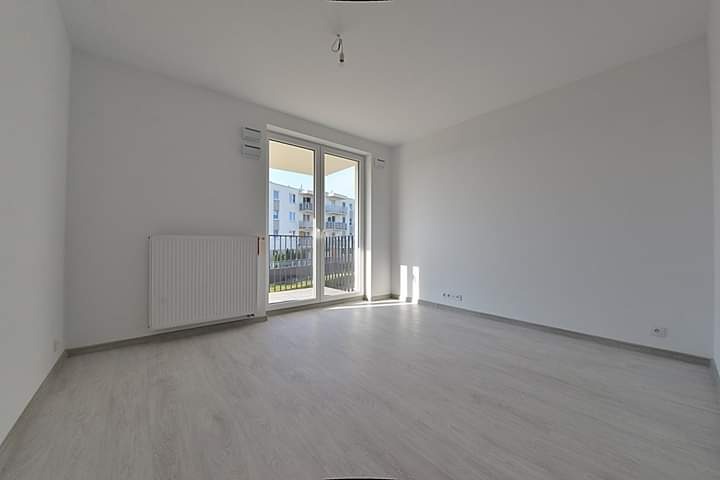 Puste, jasne pomieszczenie z panelami podłogowymi w odcieniach szarości, grzejnikiem przy ścianie i balkonem z widokiem na budynek wielorodzinny.