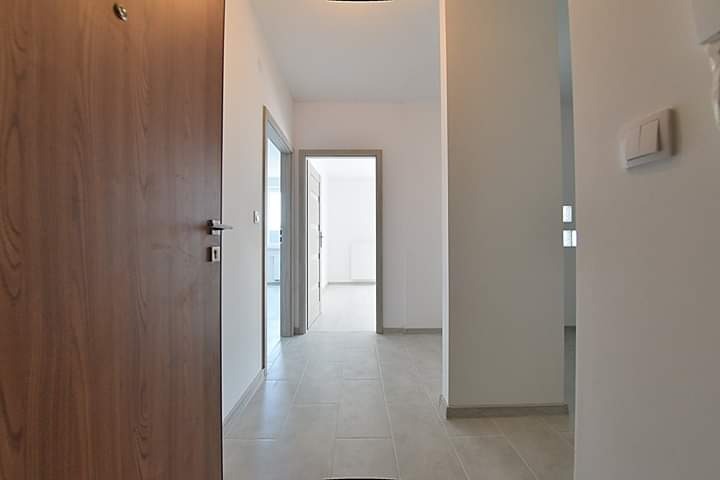 Nowoczesny korytarz z drewnianymi drzwiami wejściowymi, szarą podłogą z płytek i białymi ścianami, widok na wnętrze mieszkania przez otwarte drzwi.