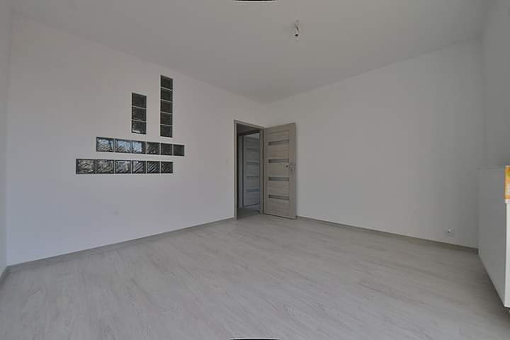 Pusty, jasny pokój z panelami podłogowymi, białymi ścianami i dekoracyjnym układem szklanych pustaków na ścianie, częściowo otwarte drzwi do sąsiedniego pomieszczenia.