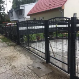 Nowoczesne, czarne ogrodzenie panelowe z bramą wjazdową, widok z zewnątrz na posesję z kostką brukową i budynkami w tle.