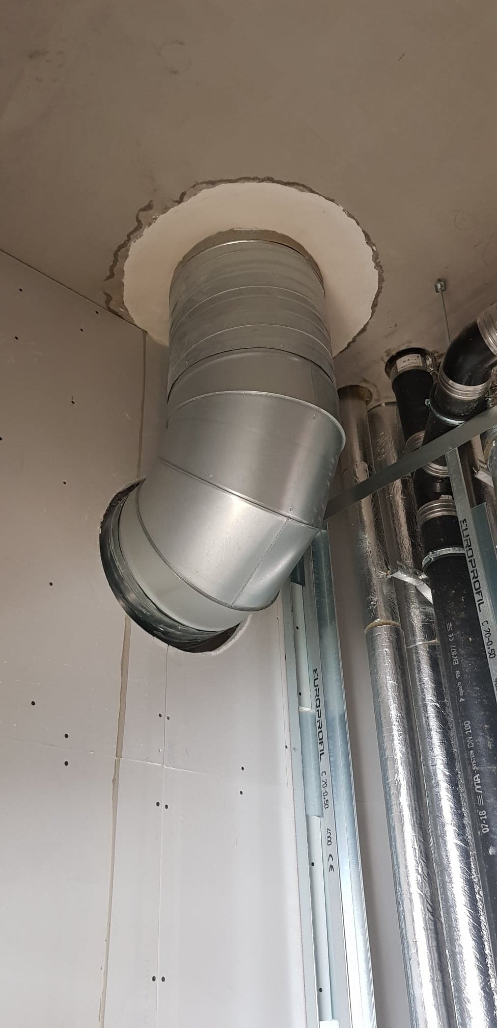 Montaż spiralnego kanału wentylacyjnego z blachy ocynkowanej, w suficie podwieszanym z płyt gipsowo-kartonowych, widoczne elementy konstrukcji nośnej sufitu i inne rury.