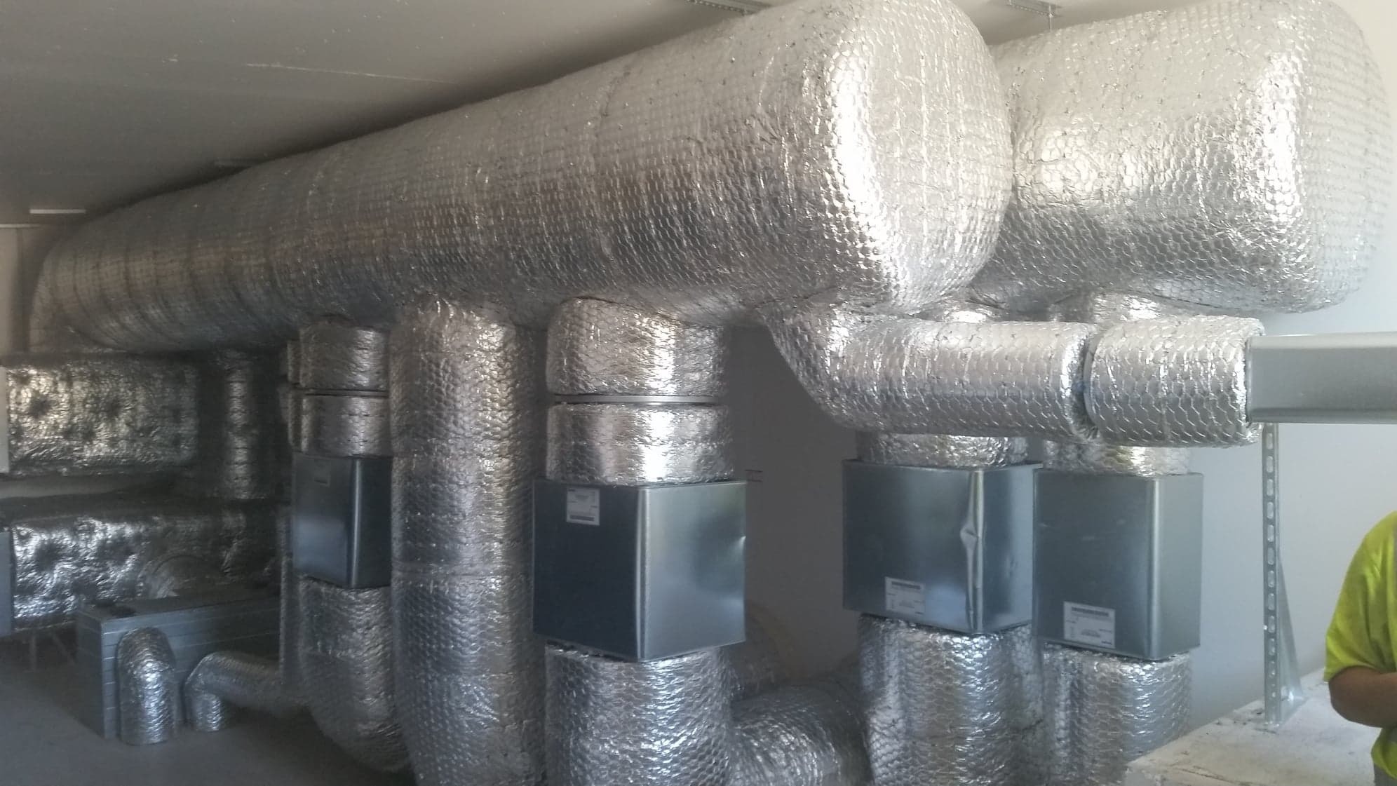 Izolowane rury wentylacyjne z aluminiową folią, widoczne czarne skrzynki na rurach, fragment osoby w jaskrawożółtej koszulce po prawej stronie kadru.