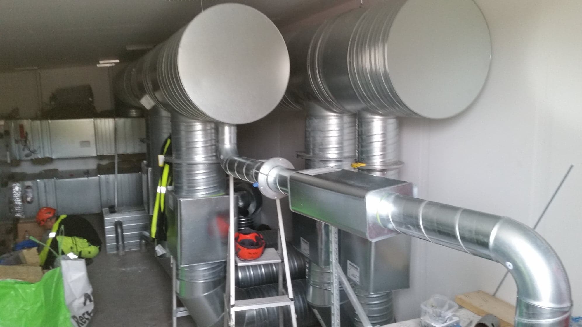 Wnętrze pomieszczenia z zamontowanymi, ocynkowanymi kanałami wentylacyjnymi o okrągłym i prostokątnym przekroju, widoczna drabina, kask ochronny i odzież robocza.