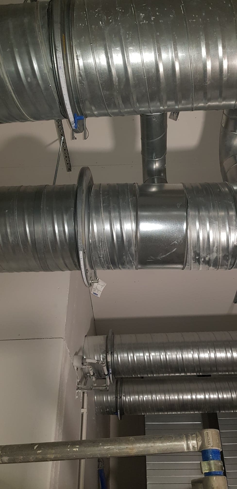 Widok instalacji wentylacyjnej z metalowych, karbowanych rur pod sufitem, z widoczną miarką zwiniętą wokół jednej z rur.