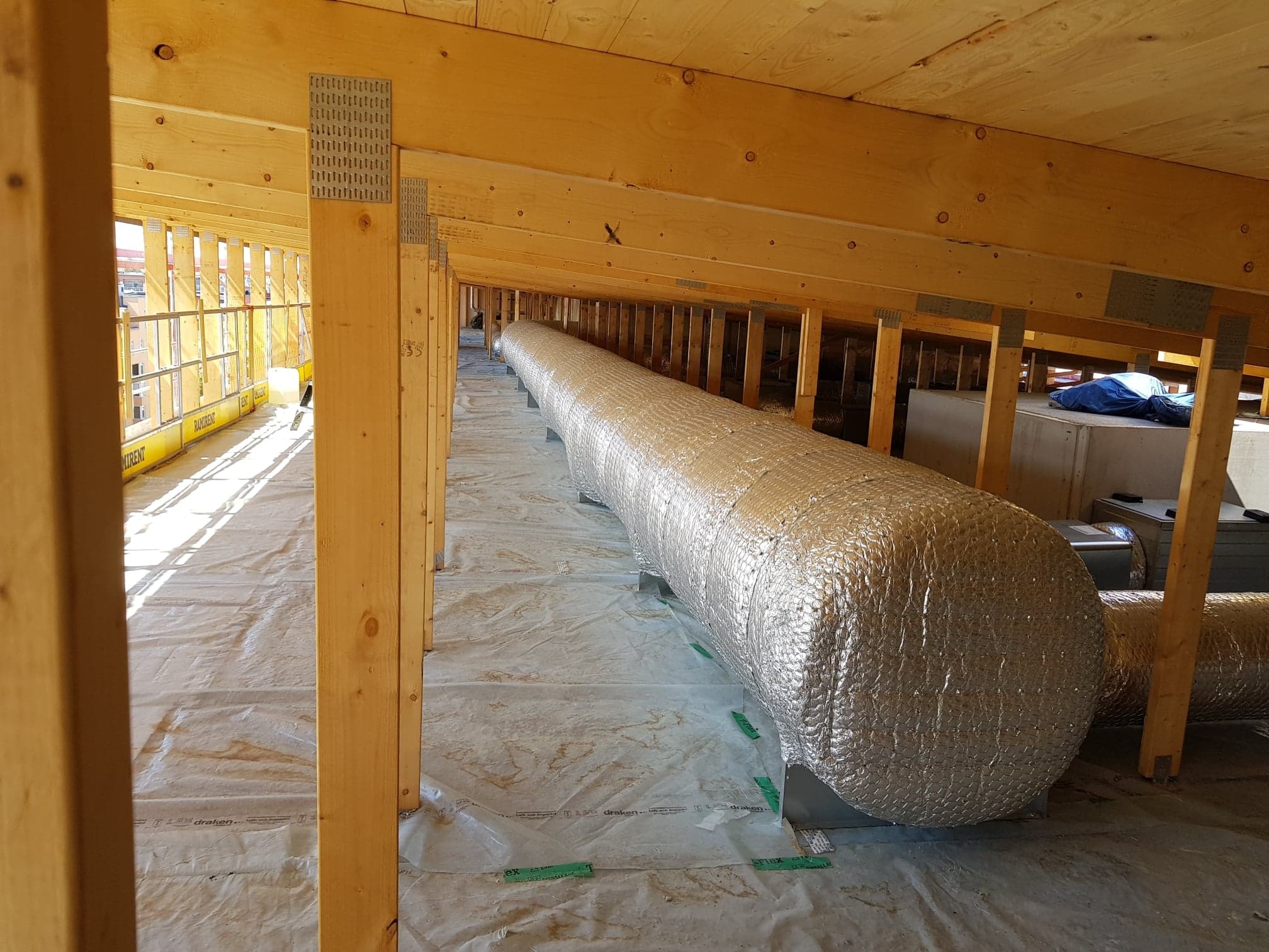 Długi, izolowany przewód wentylacyjny ułożony w przestrzeni poddasza z drewnianą konstrukcją nośną i folią ochronną na podłodze.