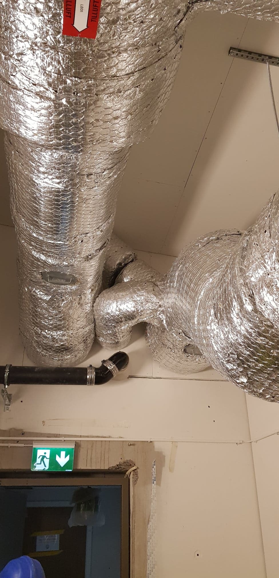 Srebrne, izolowane przewody wentylacyjne pod sufitem w budynku, z widocznym fragmentem instalacji kanalizacyjnej i znakiem ewakuacyjnym nad drzwiami.