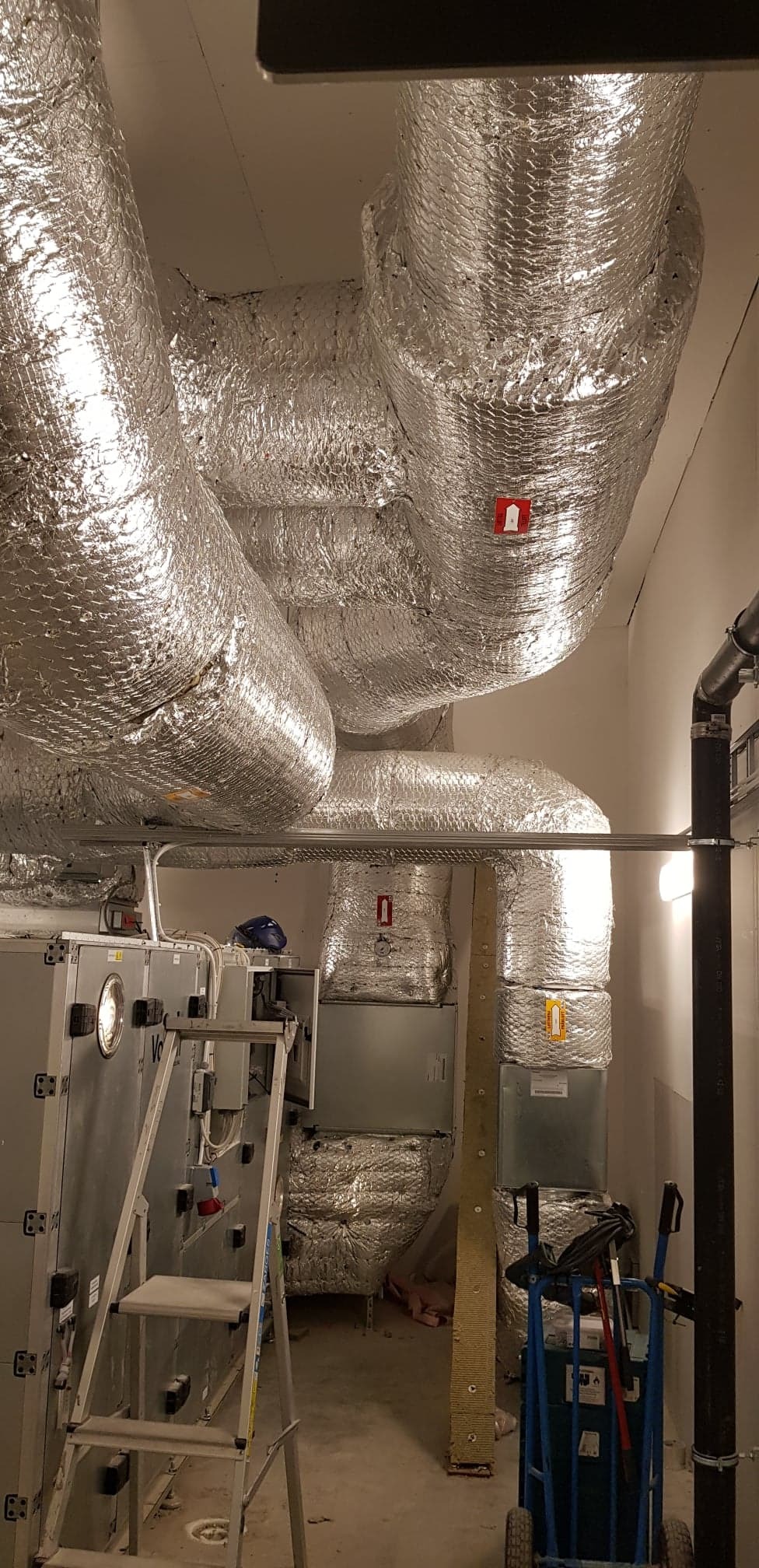 Srebrne, izolowane kanały wentylacyjne pod sufitem, widoczna drabina, wózek narzędziowy i urządzenia w pomieszczeniu technicznym.