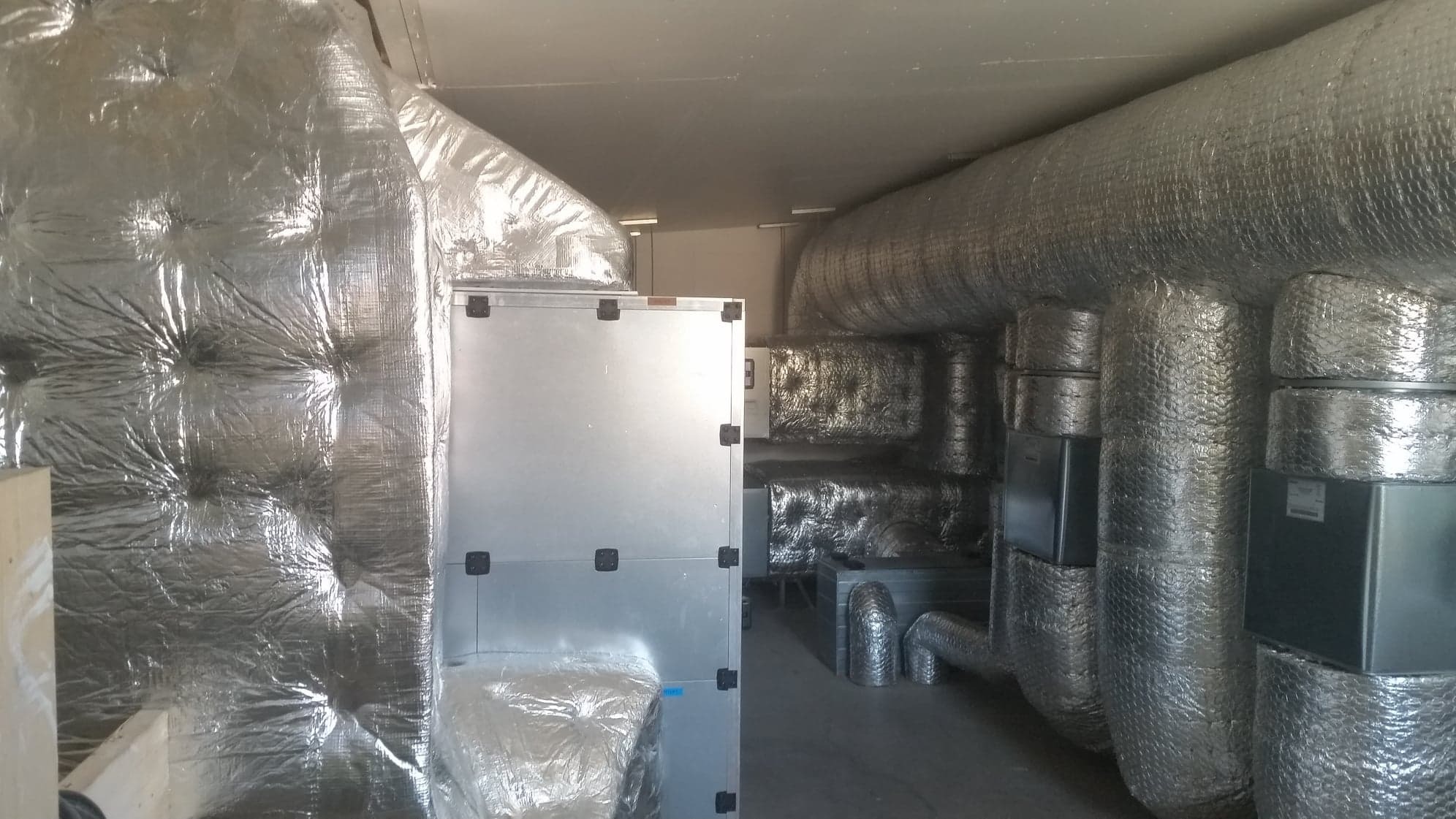 Instalacja przemysłowa: metalowe kanały wentylacyjne owinięte folią izolacyjną, widoczny centralny moduł wentylacyjny z czarnymi zatrzaskami w hali przemysłowej.