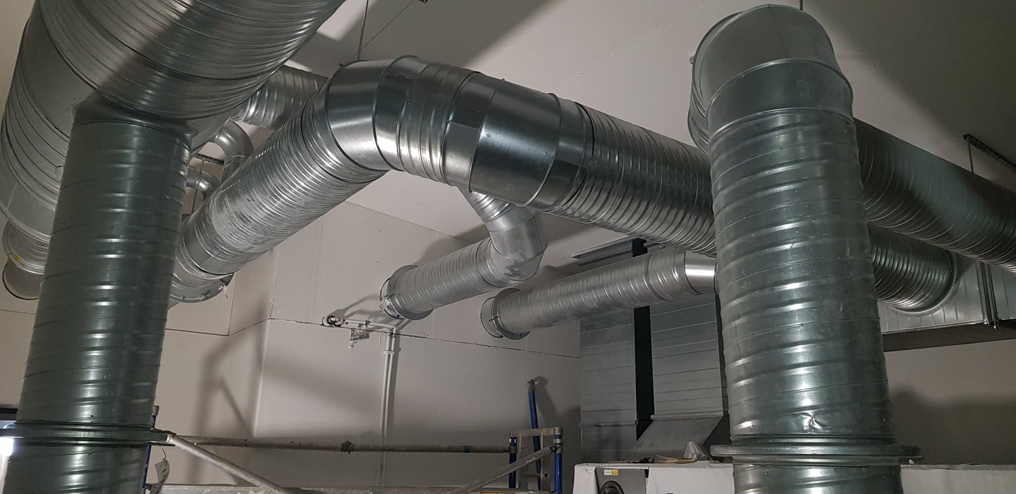 Instalacja wentylacyjna z rurami spiro, widoczne łączenia i mocowania do sufitu, częściowo zamontowana na białej ścianie w budynku komercyjnym.