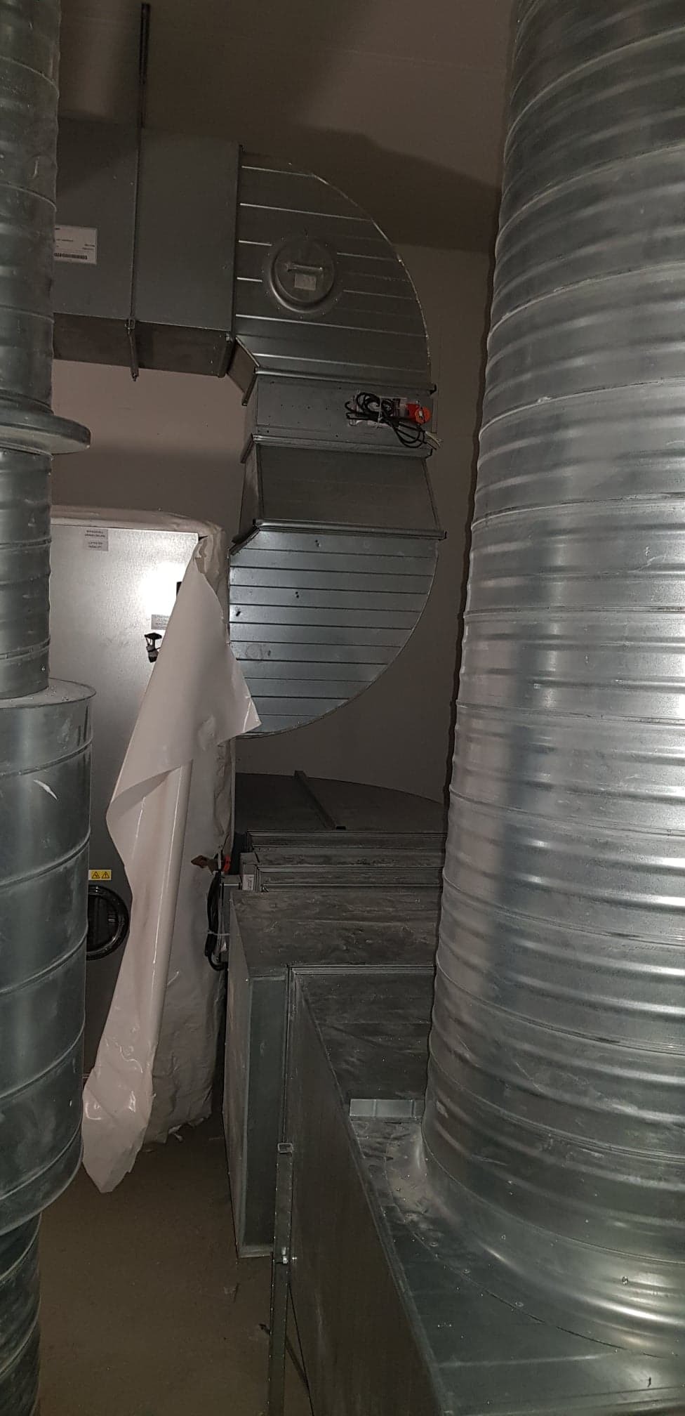 Metalowe kanały wentylacyjne, rury spiro i elementy instalacji w szarej tonacji, widoczne elementy elektryczne i zabezpieczenia.