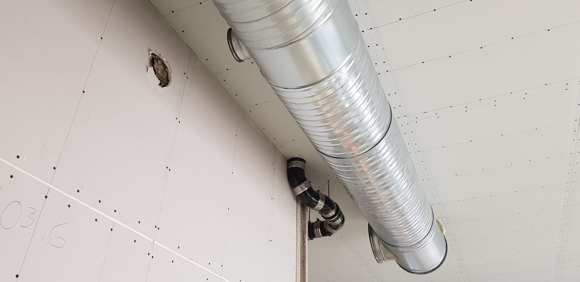 Szczegół instalacji: rura wentylacyjna z metalu falistego, połączona czarnymi rurami z tworzywa, zamontowana pod sufitem z płyt gipsowo-kartonowych, widoczne ślady po wierceniu i otwór w ścianie.