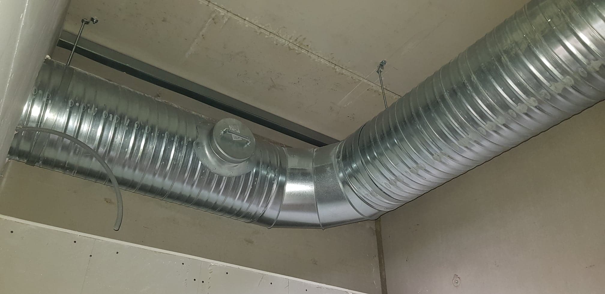 Kątowe ujęcie ocynkowanego kanału wentylacyjnego z klapą rewizyjną zamontowanego pod stropem w surowym pomieszczeniu.
