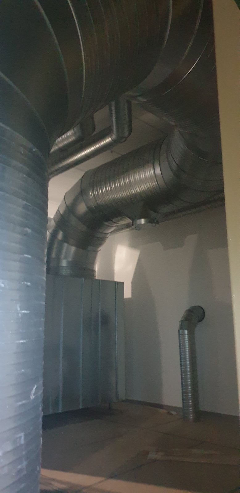 Metalowe rury wentylacyjne, częściowo izolowane, zamontowane pod sufitem w pomieszczeniu z szarą ścianą i widoczną podłogą.