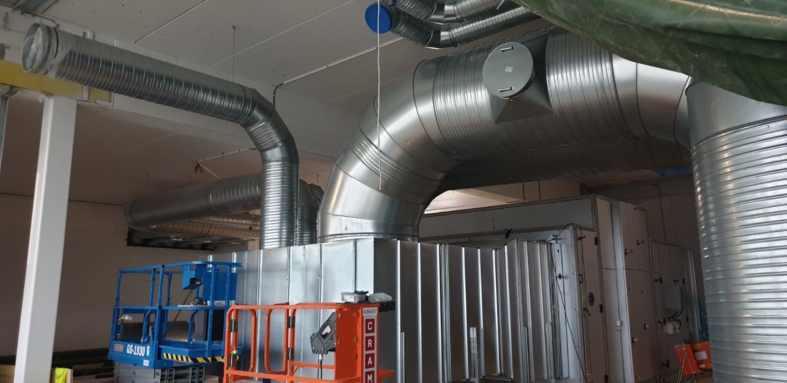 Srebrne, spiralne rury wentylacyjne zamontowane pod sufitem w budynku komercyjnym, widoczne podnośniki nożycowe w kolorach niebieskim i pomarańczowym.