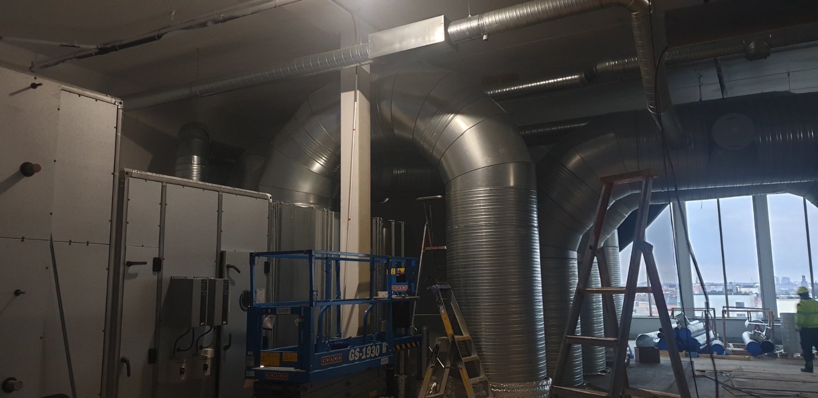Wnętrze budynku komercyjnego w trakcie instalacji systemu wentylacji z widocznymi przewodami wentylacyjnymi, platformą roboczą i drabinami.