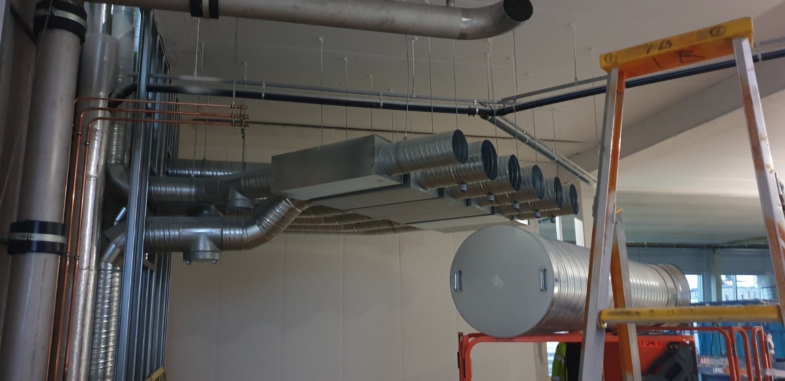 Szare rury wentylacyjne i miedziane rurki zamontowane pod sufitem w budynku komercyjnym, widoczne rusztowanie z pomarańczowymi elementami.