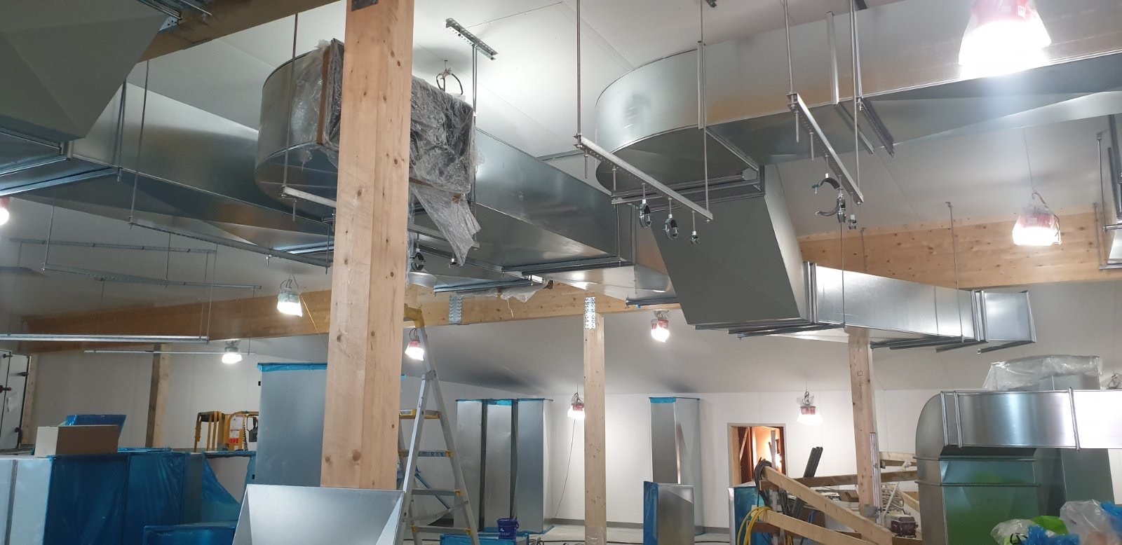 Montaż kanałów wentylacyjnych z blachy ocynkowanej w nowym budynku, widoczne drewniane słupy konstrukcyjne i drabina, niektóre elementy jeszcze w folii ochronnej.