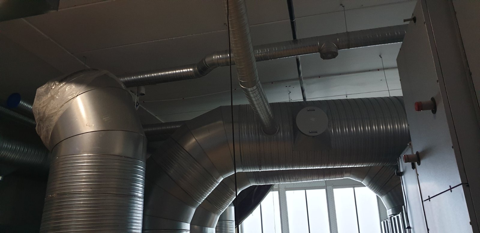 System rur wentylacyjnych z blachy ocynkowanej, widoczne elementy izolacji na jednym z kolanek, zamontowany w pomieszczeniu komercyjnym z oknami.