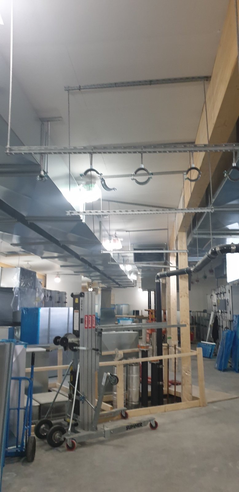 Wnętrze hali w trakcie montażu instalacji wentylacyjnej, widoczne elementy systemu podwieszone pod sufitem, kanały wentylacyjne oraz podnośnik do montażu.