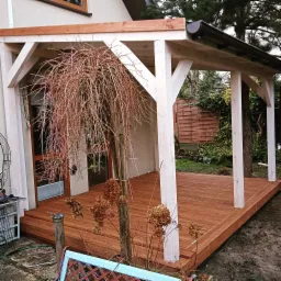 Zadaszony taras drewniany z białymi słupami, brązową podłogą i metalową rynną, ozdobiony uschniętą roślinnością i elementami dekoracyjnymi w ogrodzie.