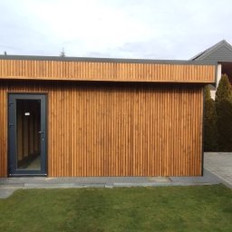 DrewKris Krzysztof Granat - Nowoczesna, drewniana konstrukcja z ciemną stolarką drzwiową, otoczona zielenią. Widoczne pionowe deski elewacyjne i starannie wykończone detale.