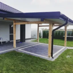 Zadaszona konstrukcja z drewnianymi słupami i czarnym dachem, przylegająca do domu z szarym brukowanym tarasem i zielonym trawnikiem.