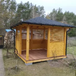 Żółta, drewniana altana z poziomymi listwami, kryta czarną dachówką bitumiczną, na fundamencie z betonowych bloczków, z drewnianą podłogą, widok z zewnątrz, na trawniku, w tle drzewa.