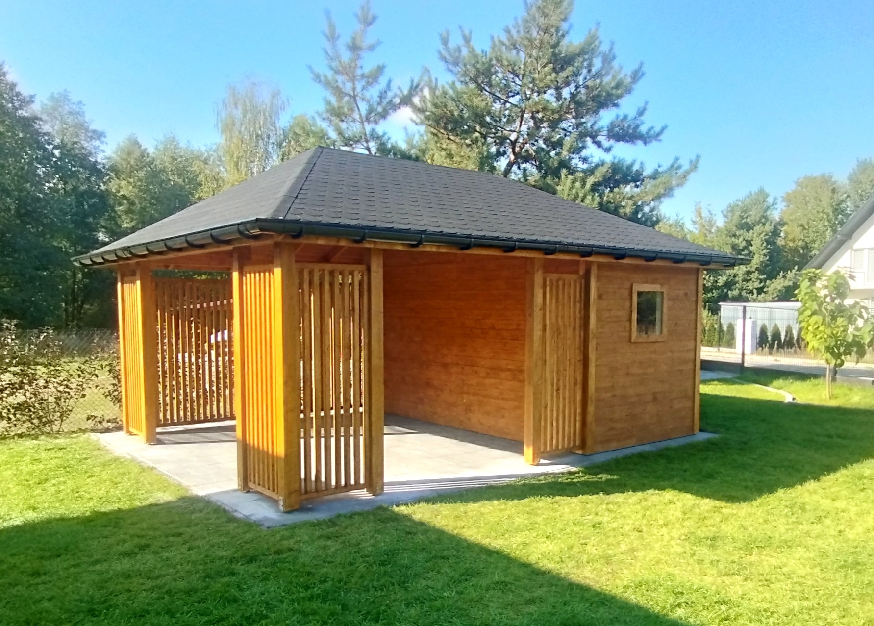 Drewniana altana z ciemnym dachem na trawniku, z widocznymi pionowymi panelami i małym oknem, otoczona zielenią w słoneczny dzień.