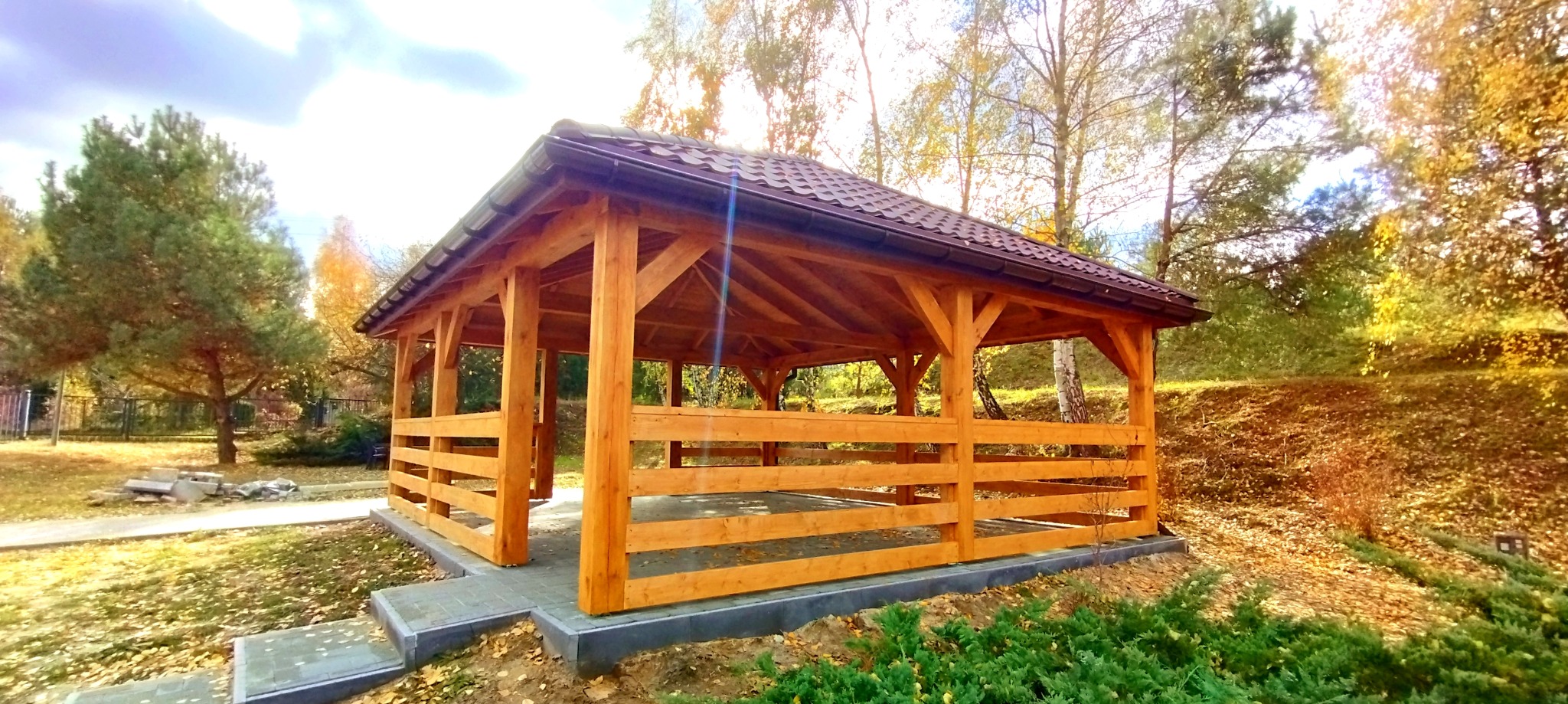 Drewniana altana z brązowym dachem na tle jesiennego krajobrazu. Konstrukcja z belek, częściowo obudowana. Widoczne schody wejściowe z szarej kostki.