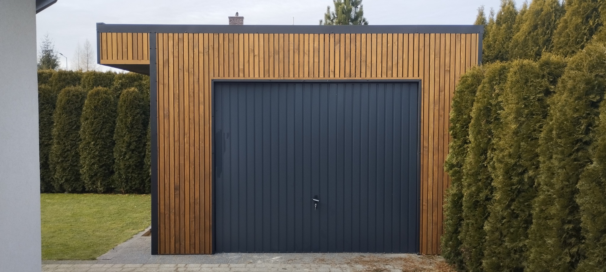 Nowoczesna, drewniana konstrukcja z garażem o grafitowych drzwiach, otoczona tujami. Widoczne pionowe deski i minimalistyczny design.